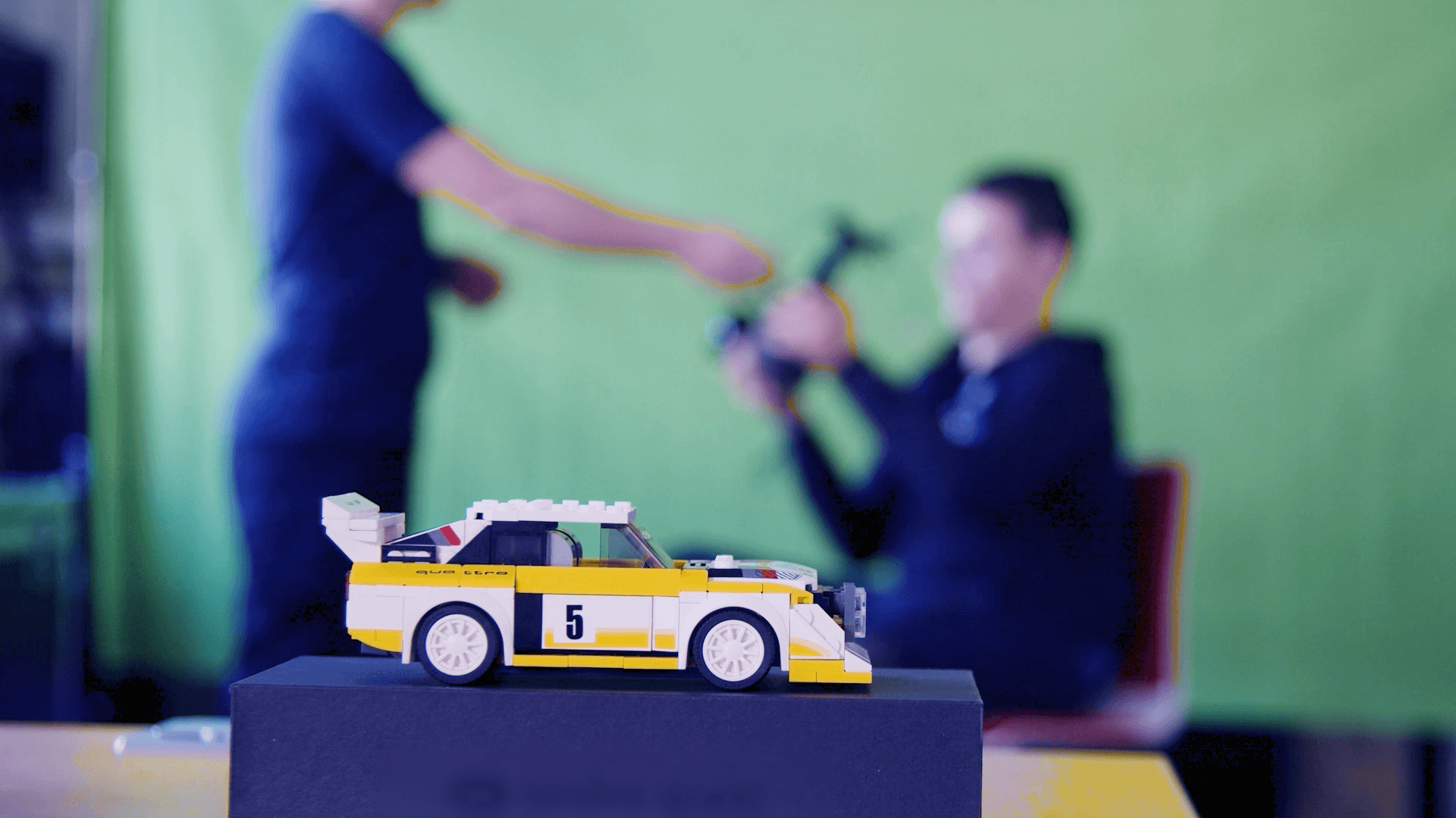 Ein gelb-weißes Legoauto im Vordergrund, dahinter verschwommen zwei Personen vor einem Greenscreen.